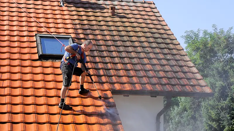 A roof cleaner soft washing a tile roof, half cleaned and half still dark with buildup